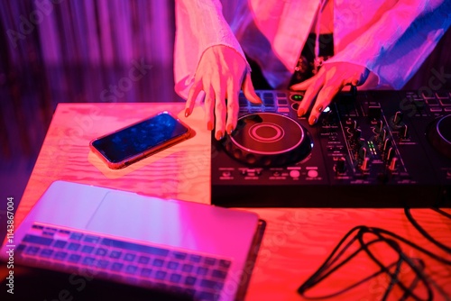 DJ’s hands operate a professional mixer on a wooden table, with a smartphone and laptop nearby, under vibrant pink and blue lighting in a club performance setting. photo