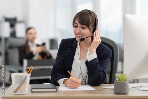 Saleswoman making notes while talking to client via headset at desk in office photo