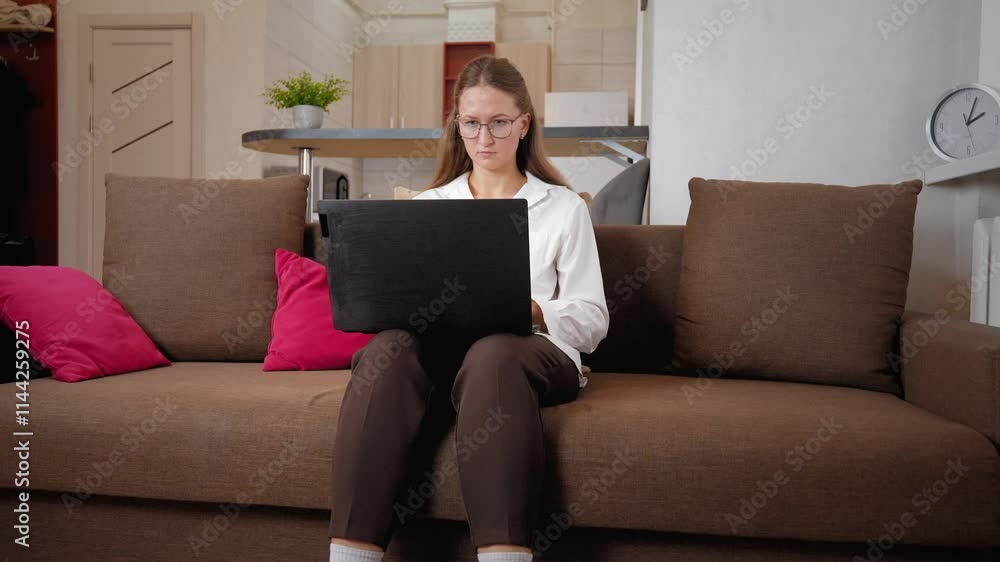 Businesswoman woman with glasses sitting on sofa in her living room and working on laptop. She wears white shirt and brown pants Businesswoman woman with glasses sitting on sofa in her living room and working on laptop. She wears white shirt and brown pants