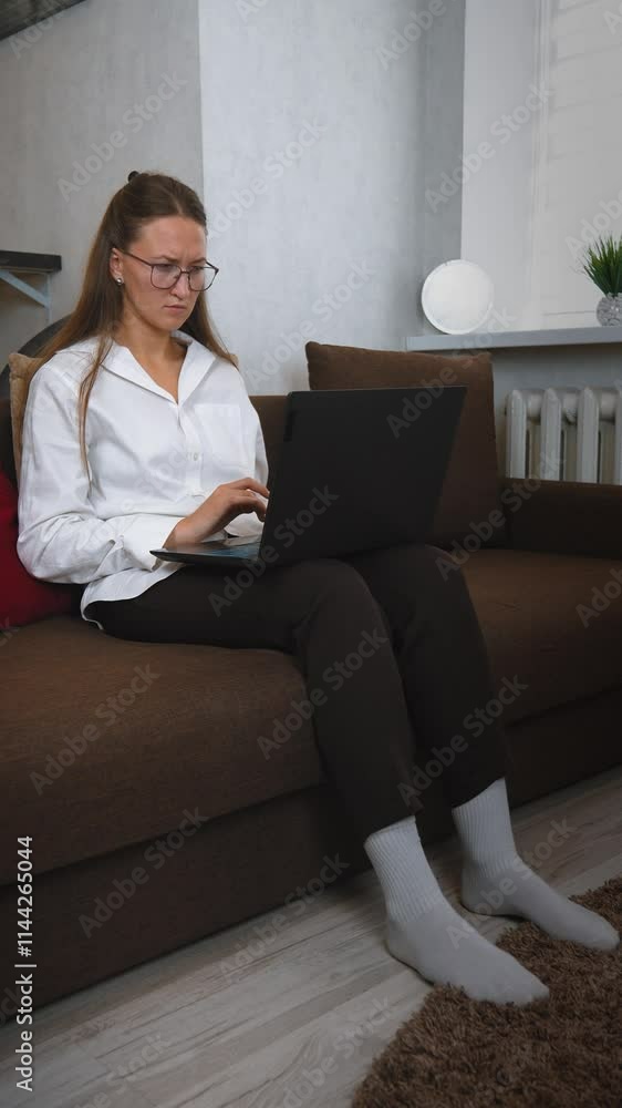 Young woman wearing glasses and business casual attire sits comfortably on brown couch, focused on typing on her laptop while working remotely in cozy home environment. vertical video Young woman wearing glasses and business casual attire sits comfortably on brown couch, focused on typing on her laptop while working remotely in cozy home environment. vertical video