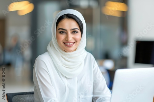 A smiling woman wearing a hijab sits at a desk with a laptop, exuding confidence and professionalism in a modern office setting.