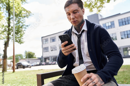 Handsome young man with tattoos sits on a bench sipping coffee and using his smartphone outdoors. photo