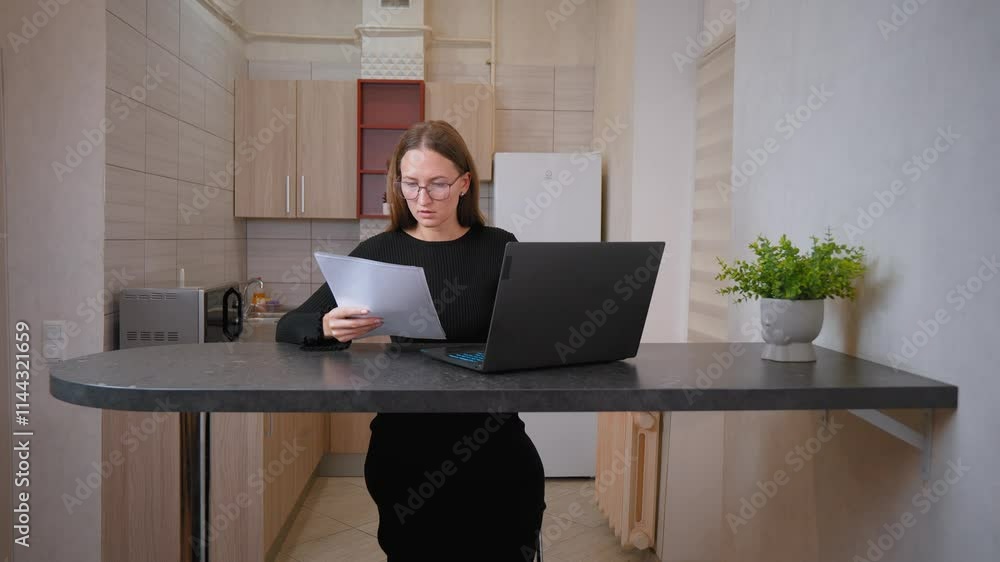 Young woman working from home looking at documents and typing on laptop. Focused and serious, she copes with her tasks in cozy atmosphere of kitchen office Young woman working from home looking at documents and typing on laptop. Focused and serious, she copes with her tasks in cozy atmosphere of kitchen office