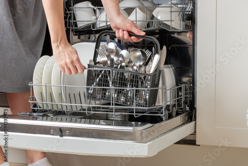 Woman's hands emptying, unloading a dishwasher, taking out a cutlery basket with spoons, knives and forks. Household chores with a modern kitchen appliance. An open dishwasher  photo