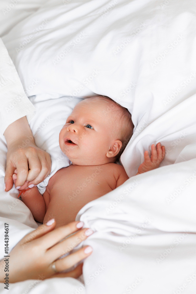 A baby is laying on a bed with its arms around a woman's hand. The baby is smiling and looking up at the camera. Concept of warmth and love between the mother and child A baby is laying on a bed with its arms around a woman's hand. The baby is smiling and looking up at the camera. Concept of warmth and love between the mother and child