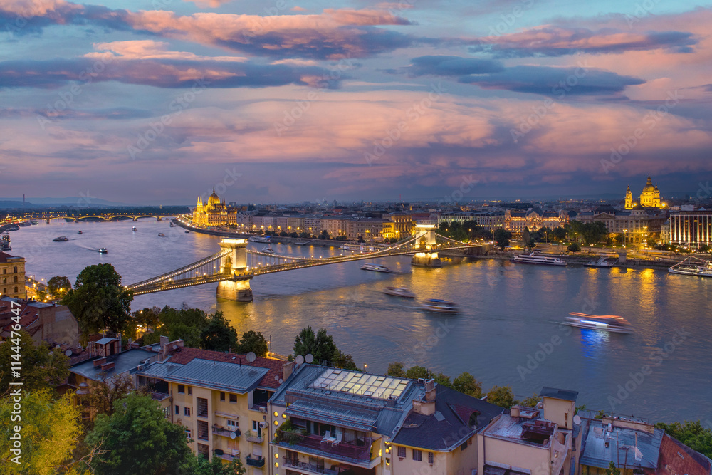 View of Budapest at dusk, residential buildings in the foreground, the Szechenyi Bridge over the Danube River, and the Pest side of the city with the Hungarian Parliament Building in the background