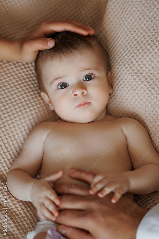 A baby is laying on a bed with a person's hand on its head. The baby has a curious expression on its face