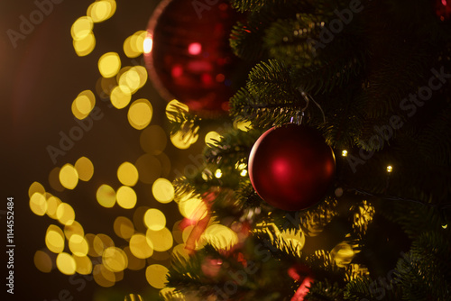 Red ornaments hang on a Christmas tree with warm lights glowing in the background during the holiday season
