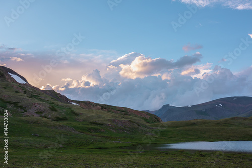 Dramatic view to small alpine lake among grassy and stone rocky hills with view to mountain range silhouette under bright golden clouds in blue sunset sky. Beautiful lake and illuminating gold sky. photo