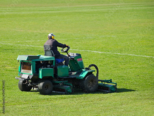 Worker on a large green lawn mower mows the grass on the football field. Landscape design and maintenance of green areas of the sports complex. Making grass stadium before the match. Human labor