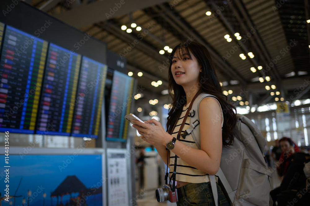 Young female tourist with backpack and camera using smartphone checking flight schedule information board in airport terminal while waiting for airplane departure Young female tourist with backpack and camera using smartphone checking flight schedule information board in airport terminal while waiting for airplane departure