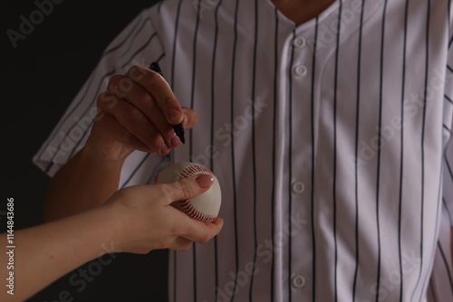 Baseball player signing autograph on ball against black background, closeup photo