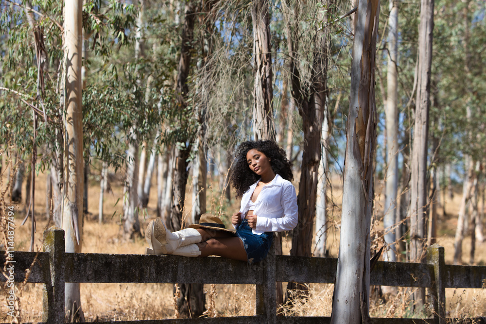 Fototapeta premium woman, young, beautiful, Latin, brunette with curly hair dressed in white shirt, denim skirt, white boots and hat, sitting with her legs stretched out on the forest fence. The woman rests.