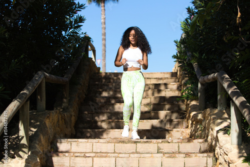 Woman, young, beautiful, Latin, brunette with curly hair, dressed in sportswear, listening to music with headphones while jogging down a staircase in the park. health and sport concept. photo