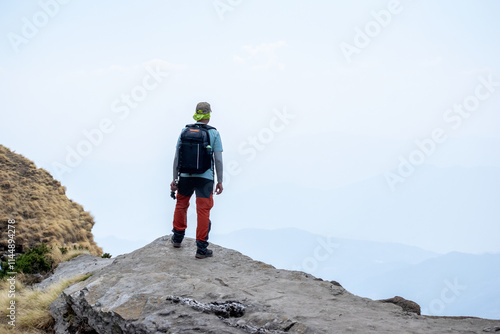 A trekker in green bandana, light blue shirt, orange pants stands atop a rocky mountain peak with a backpack, gazing into the misty horizon. Dry grassy slopes and faint mountain ridges are visible.