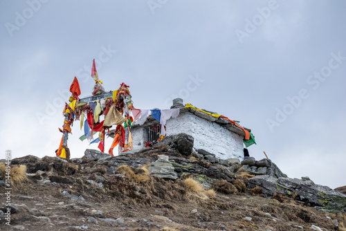 A small white stone temple perched on a rocky hilltop at chandrashila peak, adorned with colorful prayer flags and vibrant cloth offerings fluttering in the wind against a cloudy sky in Uttarakhand.
