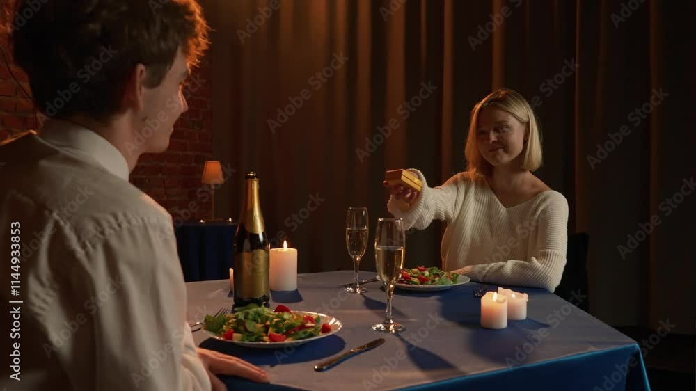 Couple celebrating valentines day and having a romantic dinner in restaurant, sitting at table and talking, woman giving to man present box, guy is surprised and happy.