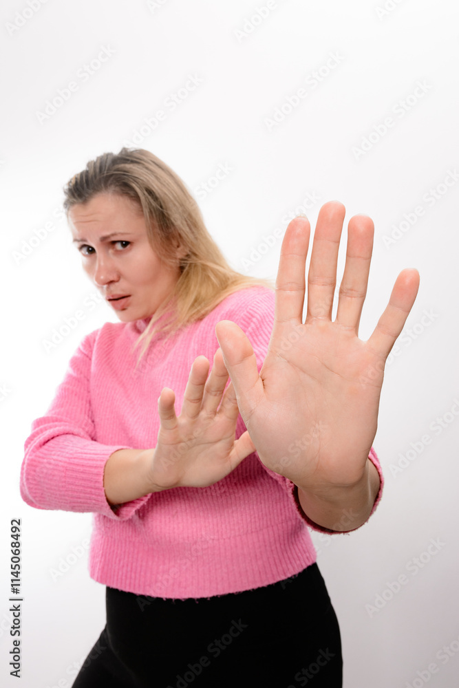 A woman with blonde hair, wearing a pink sweater and dark pants, extends her hands forward in a clear stop or refusal gesture against a light background. A woman with blonde hair, wearing a pink sweater and dark pants, extends her hands forward in a clear stop or refusal gesture against a light background.