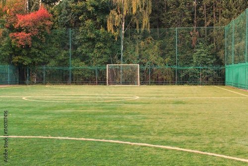 An empty soccer field artificial turf surrounded by green fence and trees. Soccer field surrounded by lush greenery. The turf with goal posts stand tall against a backdrop of vibrant autumn foliage photo