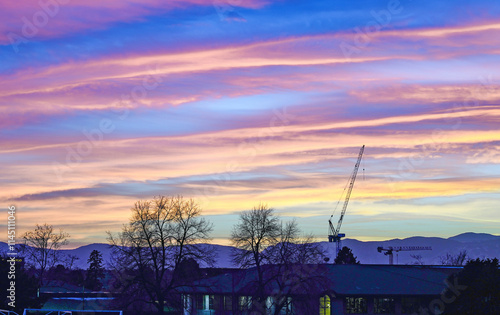 Dusk falls upon a Denver neighborhood with a colorfully striped sunset sky. A partial view of a school building in the forefront and a tall crane and mountains are visible in the background.