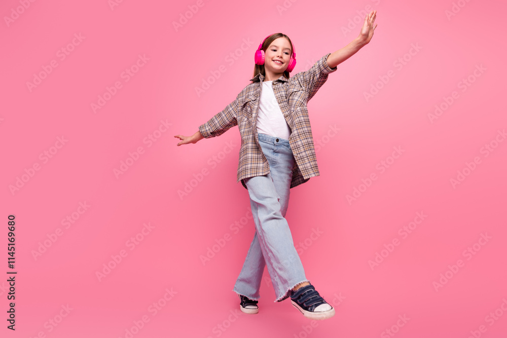 Obraz premium Young girl smiling and posing against pink background while enjoying music with headphones in a trendy casual outfit.