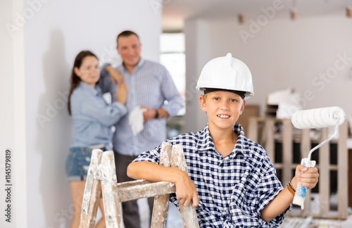 Portrait of positive young boy wearing hardhat, holding paint roller and looking at camera, standing at stepladder in apartment during repair works. His parents standing in background. photo