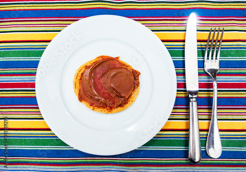 A pancake covered in dulce de leche on a large white plate with a silver knife and fork on a multicolored placemat