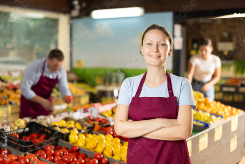 Friendly woman seller stands near garden stuff display case and gestures to invite customers to come shopping. Female store employee interrupted work and stands resting and smiling at passing visitors photo