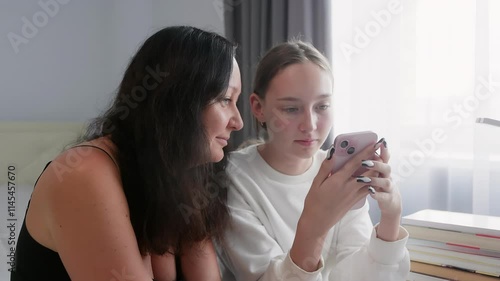 Mother and teenage daughter are using smartphone together at home, browsing internet or watching videos, sitting near the window in the bedroom