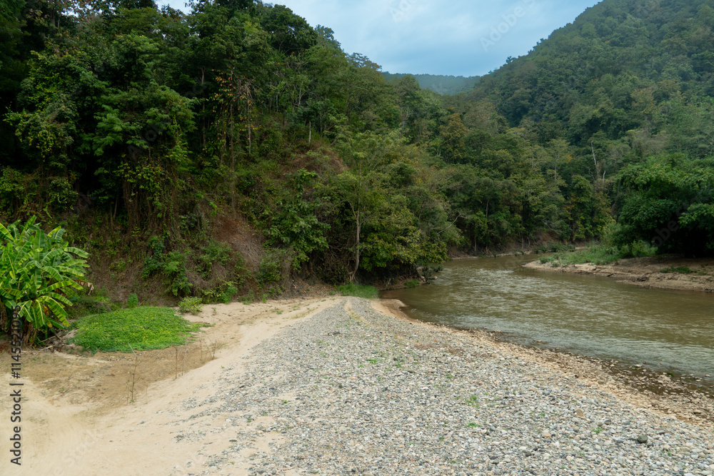 River flows through a lush green forested valley. Gravel or sandy bank lead down to the water edge. Hill or mountain covered in dense vegetation rise on either side. Mae Ngao Mae Hong Son of Thailand. River flows through a lush green forested valley. Gravel or sandy bank lead down to the water edge. Hill or mountain covered in dense vegetation rise on either side. Mae Ngao Mae Hong Son of Thailand.