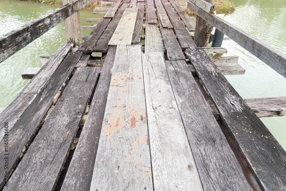 Weathered wooden bridge with some planks missing and others cracked and uneven. Spans a body of water. The water below reflects the cloudy sky and the bridge worn surface. Weathered wooden bridge with some planks missing and others cracked and uneven. Spans a body of water. The water below reflects the cloudy sky and the bridge worn surface.