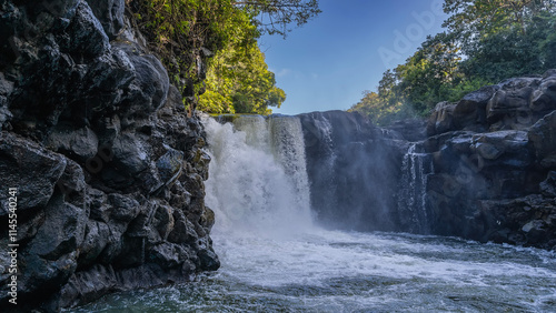 Fototapeta Naklejka Na Ścianę i Meble -  A beautiful tropical waterfall. Streams of water fall from the edge of the ledge into the river. Foam, splashes. Wet steep coastal cliffs. Green trees against a blue sky. Mauritius. GRSE waterfall