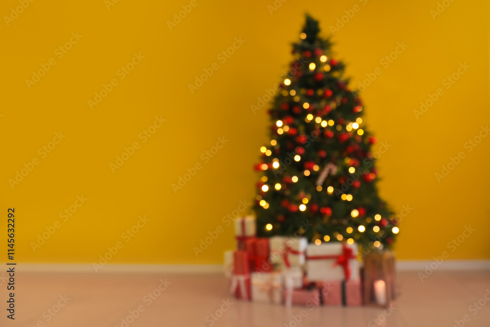 Blurred view of Christmas tree with glowing lights, ornaments and gift boxes near yellow wall Blurred view of Christmas tree with glowing lights, ornaments and gift boxes near yellow wall