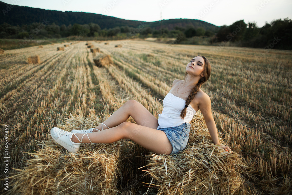 girl sitting on a haystack in a field. She is wearing a white sleeveless top and denim shorts. On her feet are white sneakers with ties. girl sitting on a haystack in a field. She is wearing a white sleeveless top and denim shorts. On her feet are white sneakers with ties.