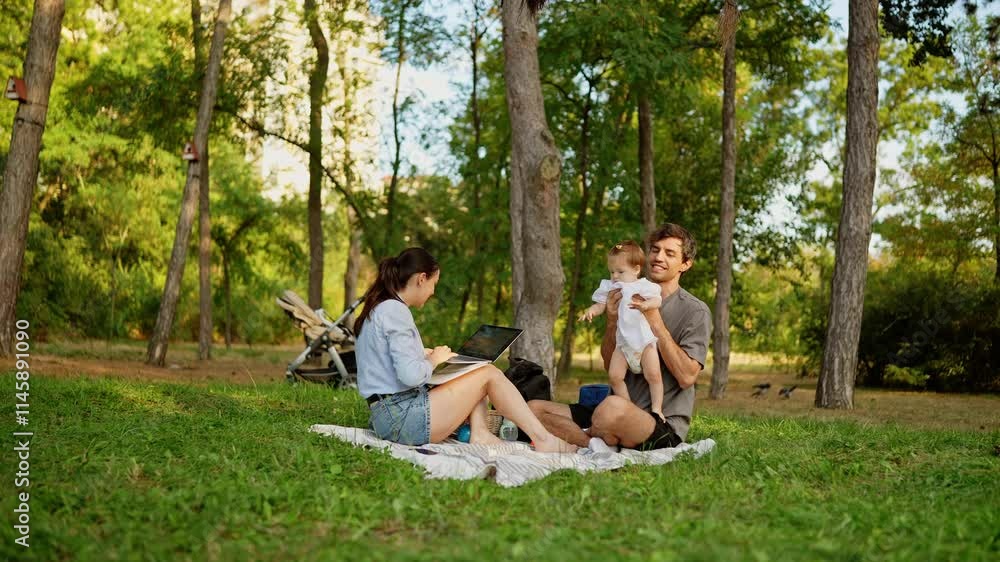 Little girl in a white dress wonders what her mother is doing on the laptop while her father takes her into his arms while relaxing in the park in summer Little girl in a white dress wonders what her mother is doing on the laptop while her father takes her into his arms while relaxing in the park in summer