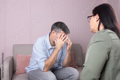 A man in therapy covers his face with his hands, showing emotion or stress, while his psychotherapist, with his back turned, listens to him in a serene environment. 