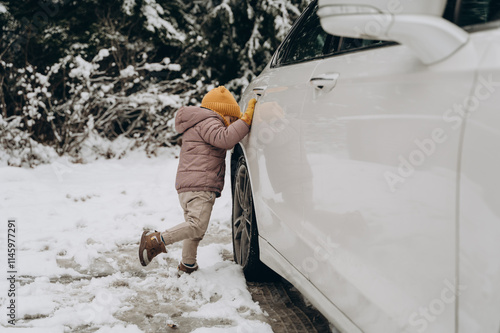 A small boy wearing a mustard cap and mustard gloves, a warm jacket and warm boots swings to hit the wheel of a white car while standing in the snow. photo