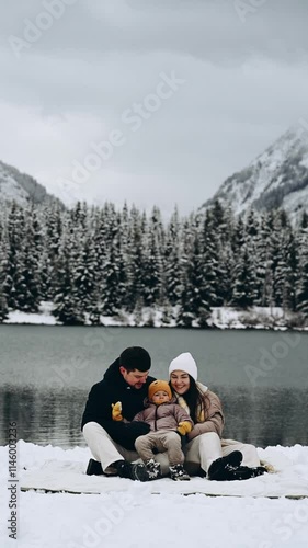 A happy family of three sits in the snow by the lake, all dressed in warm winter clothes. In the background, majestic mountains are covered with snow.