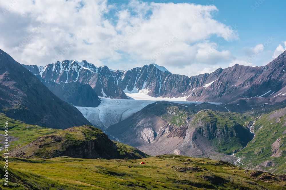 Fototapeta premium Colorful landscape with orange tent in alpine valley with view to big glacier tongue and large snow-capped mountain range under clouds in blue sky. Green hills and rocks against ice and sheer crags.