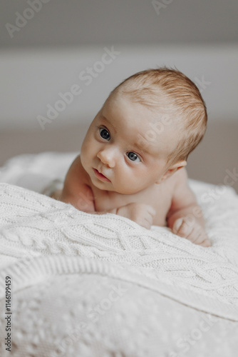 A baby is laying on a blanket with its head tilted to the side. The baby has blue eyes and is looking at the camera. Concept of innocence and curiosity