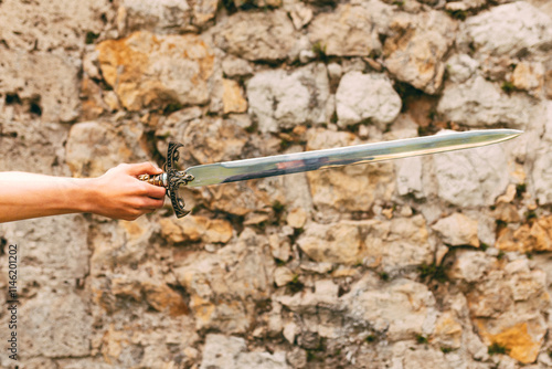 A hand holding an ornate medieval sword with a detailed hilt, set against a rustic stone wall background. Perfect for historical, fantasy, or medieval-themed concepts.