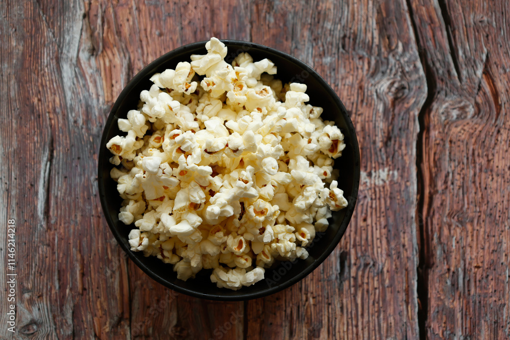 A black bowl filled with freshly popped popcorn placed on a rustic wooden table. A black bowl filled with freshly popped popcorn placed on a rustic wooden table.