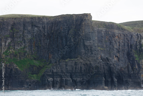 Dramatic, dark cliffs rise sharply from the ocean, layered with strata and small vegetation.  Waves crash at the base, illustrating the powerful forces of nature. photo