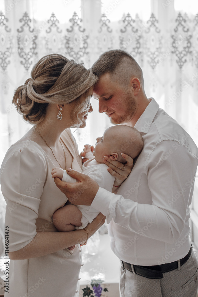Fototapeta premium A young couple holds a baby in their arms, with the baby's head resting on the man's shoulder. The woman is wearing a white dress and the man is wearing a white shirt