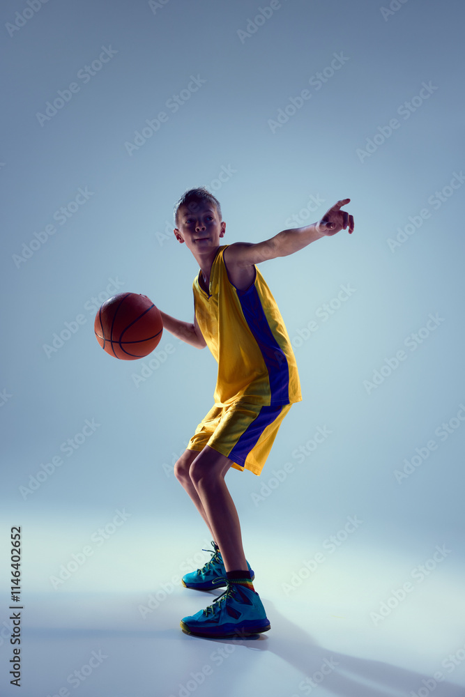 Competitive, confident child, athletic boy playing basketball against grey studio background. Junior basketball player training passing. Concept of sport, leisure activity, hobby, competition. Ad