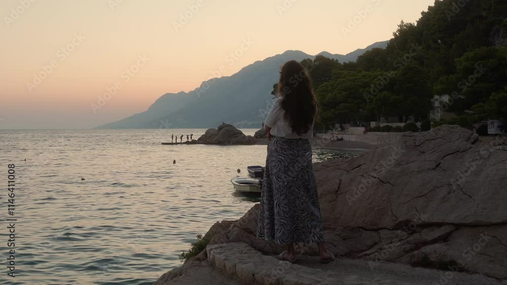 A thoughtful girl, standing alone on a stone pier in the evening twilight, dreamily looks at the sea. A woman romantically looks at the sunset on the Adriatic coast of Croatia. A thoughtful girl, standing alone on a stone pier in the evening twilight, dreamily looks at the sea. A woman romantically looks at the sunset on the Adriatic coast of Croatia.