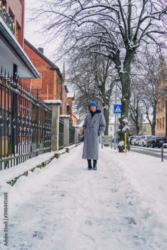 A woman stands on a snowy street, wearing a grey coat and colorful hat, capturing a serene winter moment.