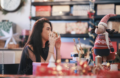 Sipping a warm beverage, a woman relishes a creative moment surrounded by colorful decorations and Christmas gifts, embracing the joyful holiday spirit photo