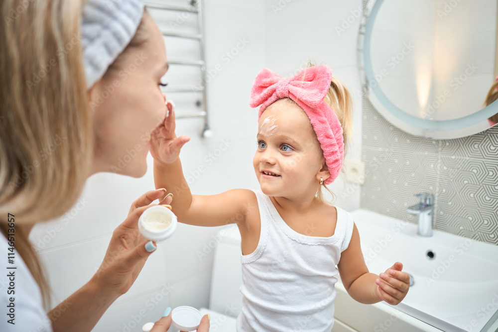 Smiling young woman and little daughter with a cosmetic bow on their heads smearing face cream on faces, during skincare procedure in the bathroom at home Smiling young woman and little daughter with a cosmetic bow on their heads smearing face cream on faces, during skincare procedure in the bathroom at home