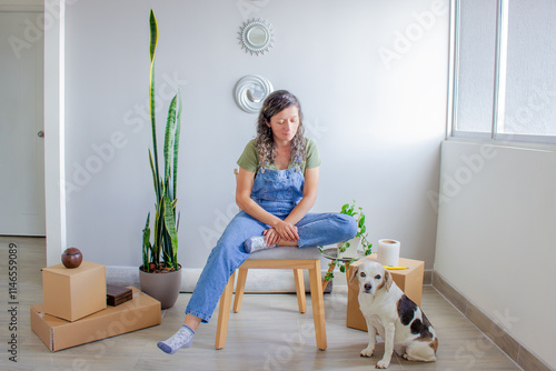 A woman sits on a chair in the middle of a room with a Beagle dog by her side. Around them are cardboard boxes, plants, and a rolled-up rug, capturing a warm moving day moment. photo